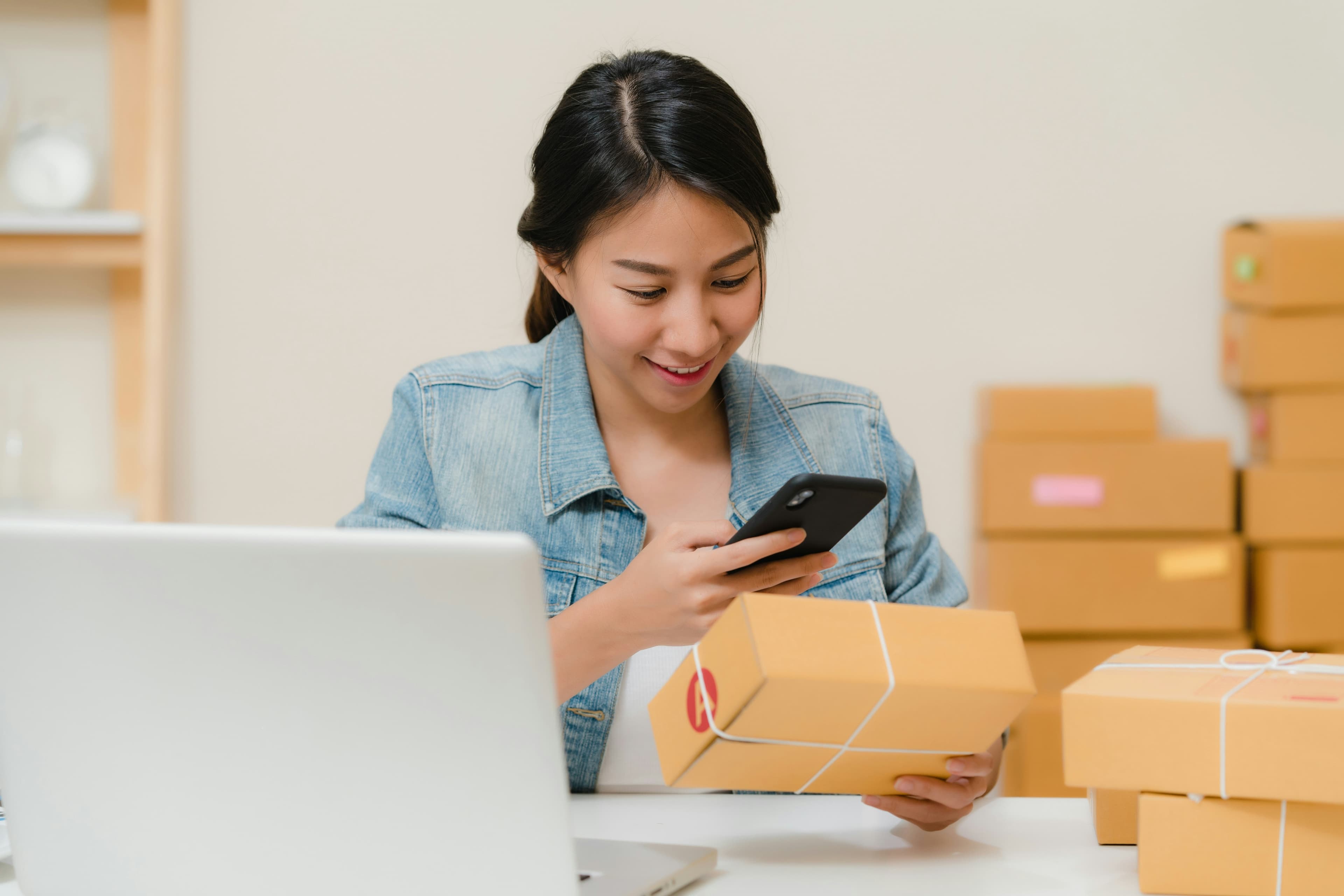 Persona revisando un paquete con el móvil junto a un portátil y cajas de envío